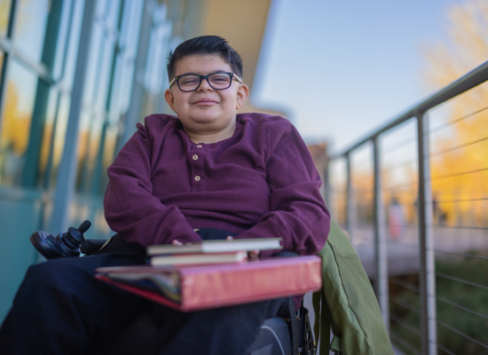 Learn about SAS Eligibility Student smiling in a wheelchair holding a pile of books.