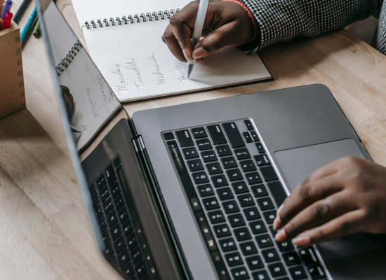 A person writes notes in a notebook beside an open laptop on a desk, with days of the week visible on the page.