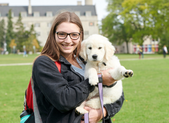 Female student wearing glasses and smiling while holding a white puppy on Johnson Green