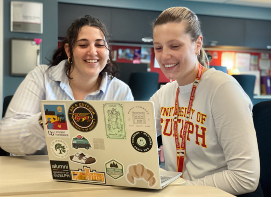 Two people sit at a table and look at a laptop covered in stickers. Both are smiling, and one wears a University of Guelph shirt and lanyard.