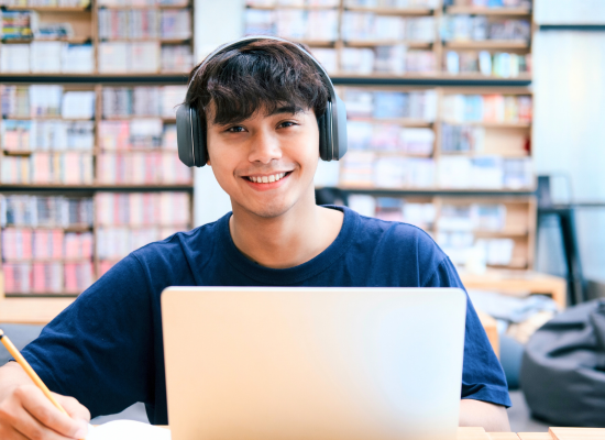 A smiling student wearing large black headphones sits in a library or study space, working on a laptop and holding a pencil, with bookshelves in the background.