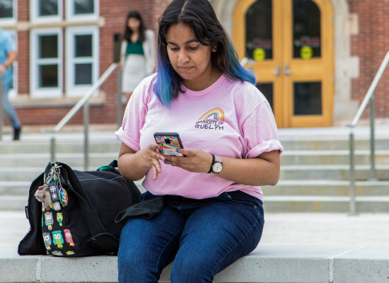 Student wearing a pink shirt sitting on a bench looking at a phone.