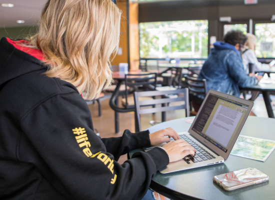 A student wearing a black hoodie works on a laptop at a round table in a brightly lit campus common area.