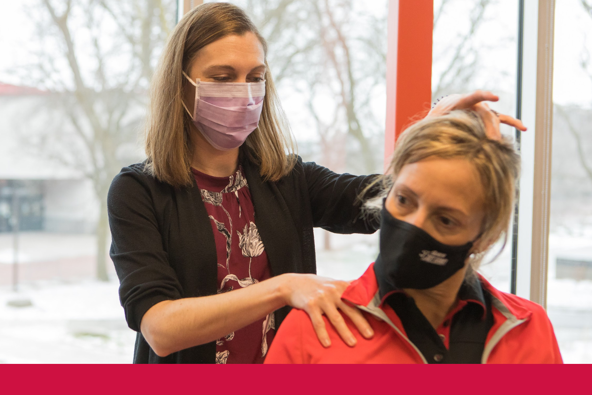 Female chiropractor performing a neck manipulation on a blonde woman wearing a black mask and red shirt