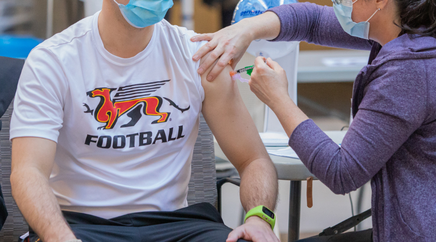 Man wearing a Gryphon football white tshirt and mask getting vaccinated by a nurse