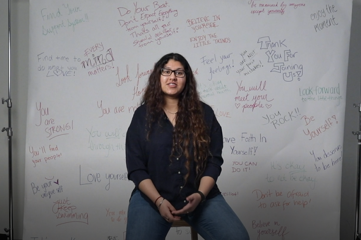 Herleena sitting on a stool in front of a white backdrop with positive affirmations written on it