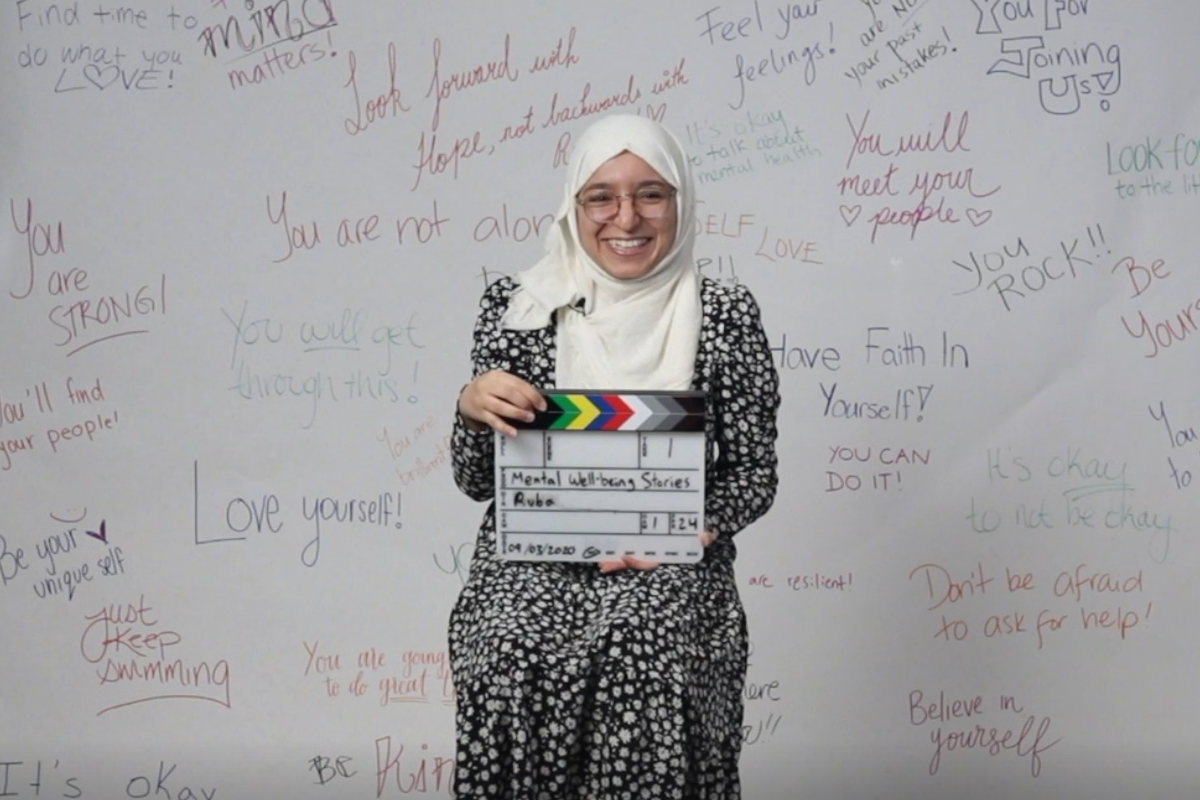 Ruba sitting on a stool holding a clapperboard in front of a white backdrop with positive affirmations written on it