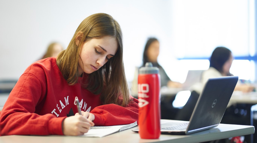 Student writing on paper at a desk with a laptop